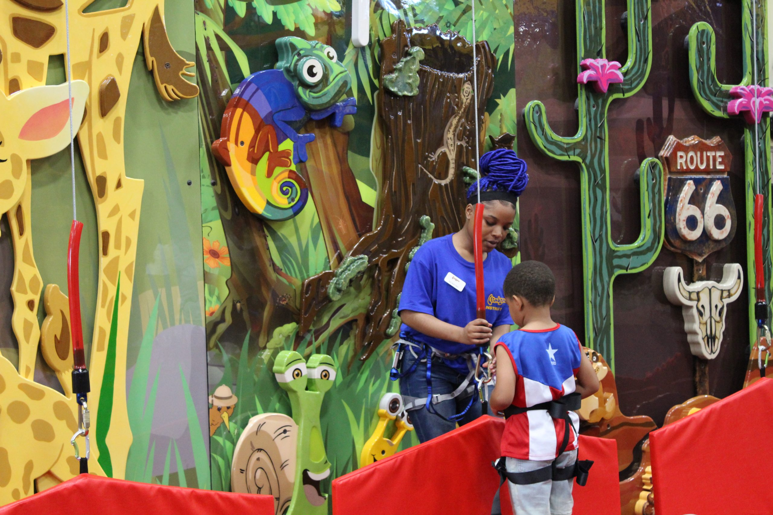 A ClimbZone instructor clipping a young climber into his harness in front of a jungle-themed wall